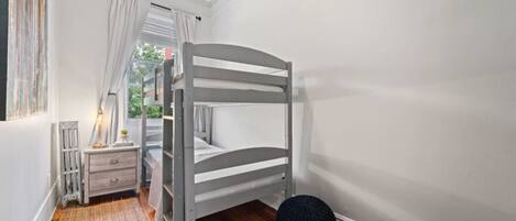 A room with a bunk bed, a small bedside table with a lamp, a red and black pouf, and a window with curtains.
