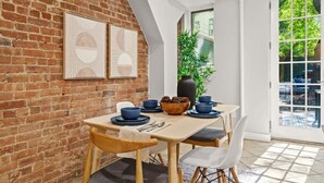 A dining area with a brick wall, two framed artworks, a wooden table, blue plates, and a potted plant.