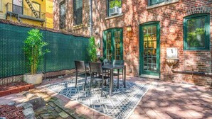 A courtyard with a patterned rug, a table and chairs, a potted plant, and a brick building with green doors and windows.