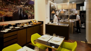 A restaurant with a dining area featuring tables set for four, a kitchen area with chefs preparing food, and a large mural on the wall depicting a market scene. - PALAZZO CARTARI (Palermo)