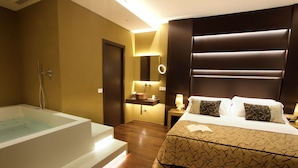 A modern hotel room with a large bathtub, a bed with patterned bedding, a bedside table with a lamp, and a mirror with a ring light. - PALAZZO CARTARI (Palermo)