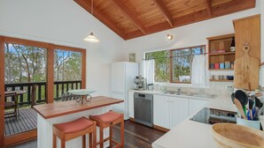 A modern kitchen with wooden cabinets, a built-in dishwasher, and a large window offering a view of trees. - 129 Murrah Street - River Views (Bermagui)