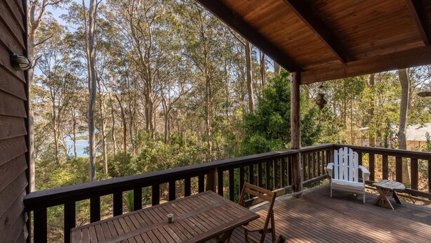 A wooden deck with a table and chairs, overlooking a forested area with a lake. - 129 Murrah Street - River Views (Bermagui)