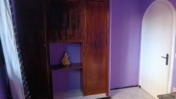 A room with a purple wall, a wooden wardrobe with a shelf, a white door, and a small table with a floral table runner.
