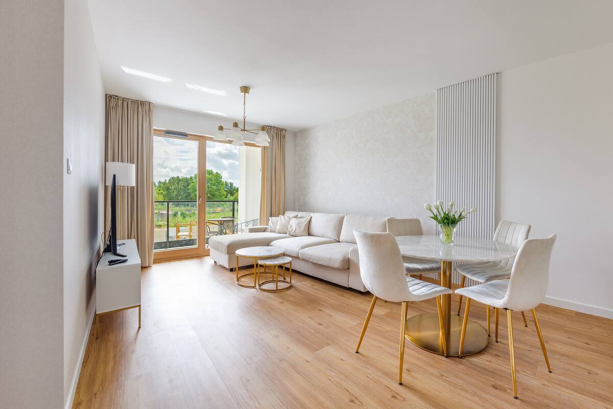 A modern living room with a white sofa, a dining table with chairs, and a balcony with a view of greenery.