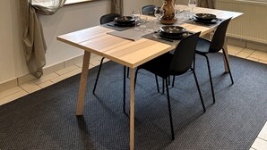 Ein Essbereich mit einem Tisch, gedeckt für vier Personen, einschließlich Tellern, Besteck und Gläsern. Zwei Stühle, eine Vase mit getrockneten Pflanzen und einer Wandleuchte sind vorhanden.