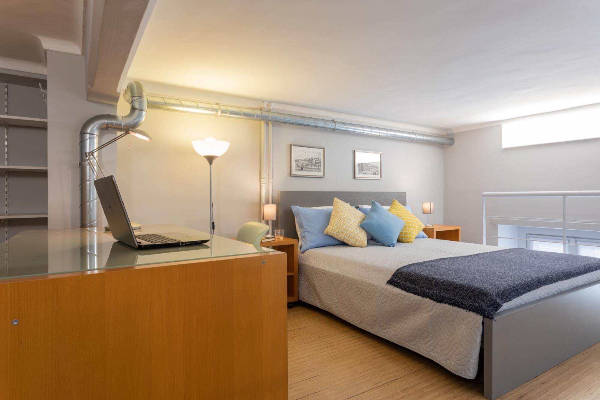 A modern bedroom with a bed, bedside tables, a desk with a laptop, and a lamp.