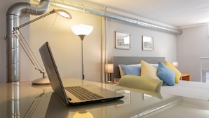A modern hotel room with a glass desk, a laptop, a bedside lamp, a bed with pillows, and a headboard.