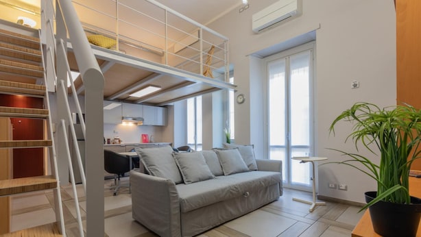 A modern living room with a grey sofa, a staircase, and a kitchen in the background.