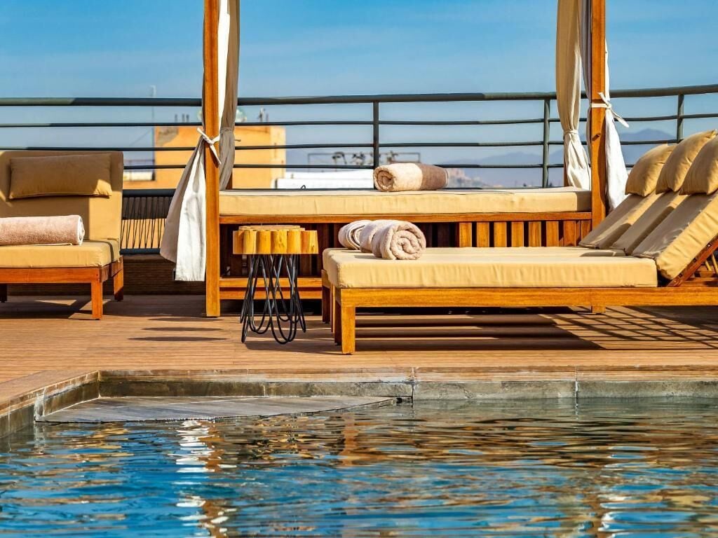 A rooftop pool area with wooden loungers, a small table, and a view of the city skyline.