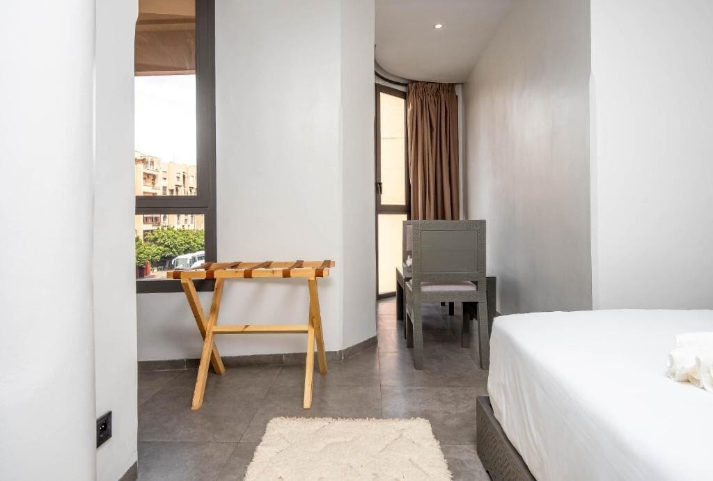 A bedroom with a bed, a wooden chair, a small table, a window with curtains, and a view of buildings outside.