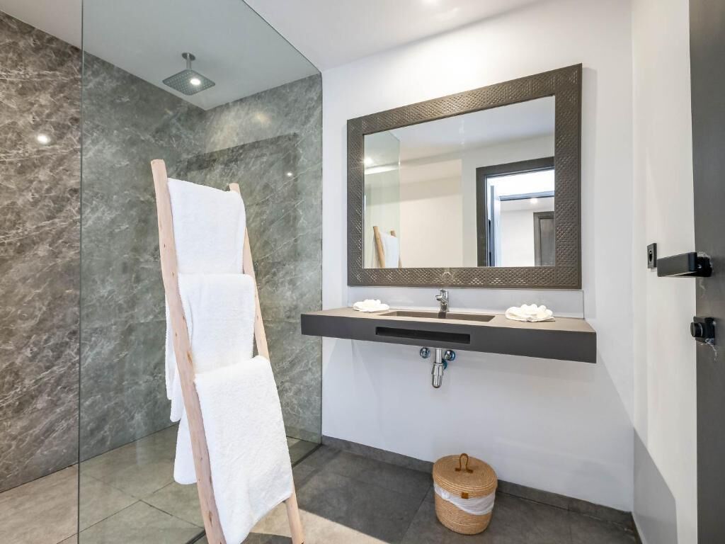 A modern bathroom with a marble wall, a wooden towel rack, a large mirror, and a sink with a towel dispenser.