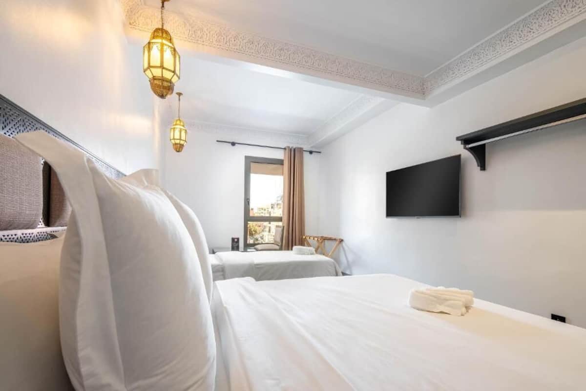 Hotellihuone, jossa on kaksi sänkyä, seinälle asennettu taulutelevisio ja suuri ikkuna, jossa on verhot.