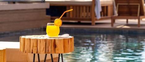 Une table en bois avec un verre de jus d’orange et une paille, placée au bord d’une piscine.
