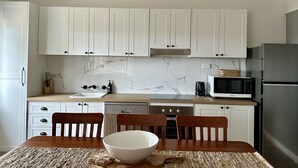 Una cocina con armarios blancos, salpicadero de mármol, una mesa de comedor de madera con sillas y un refrigerador negro.