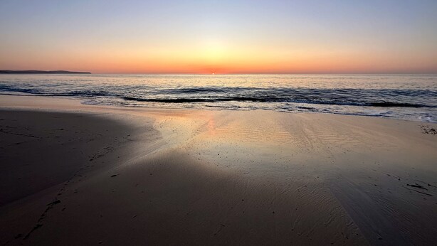 Una playa con un atardecer sobre el océano, orilla arenosa y olas suaves.