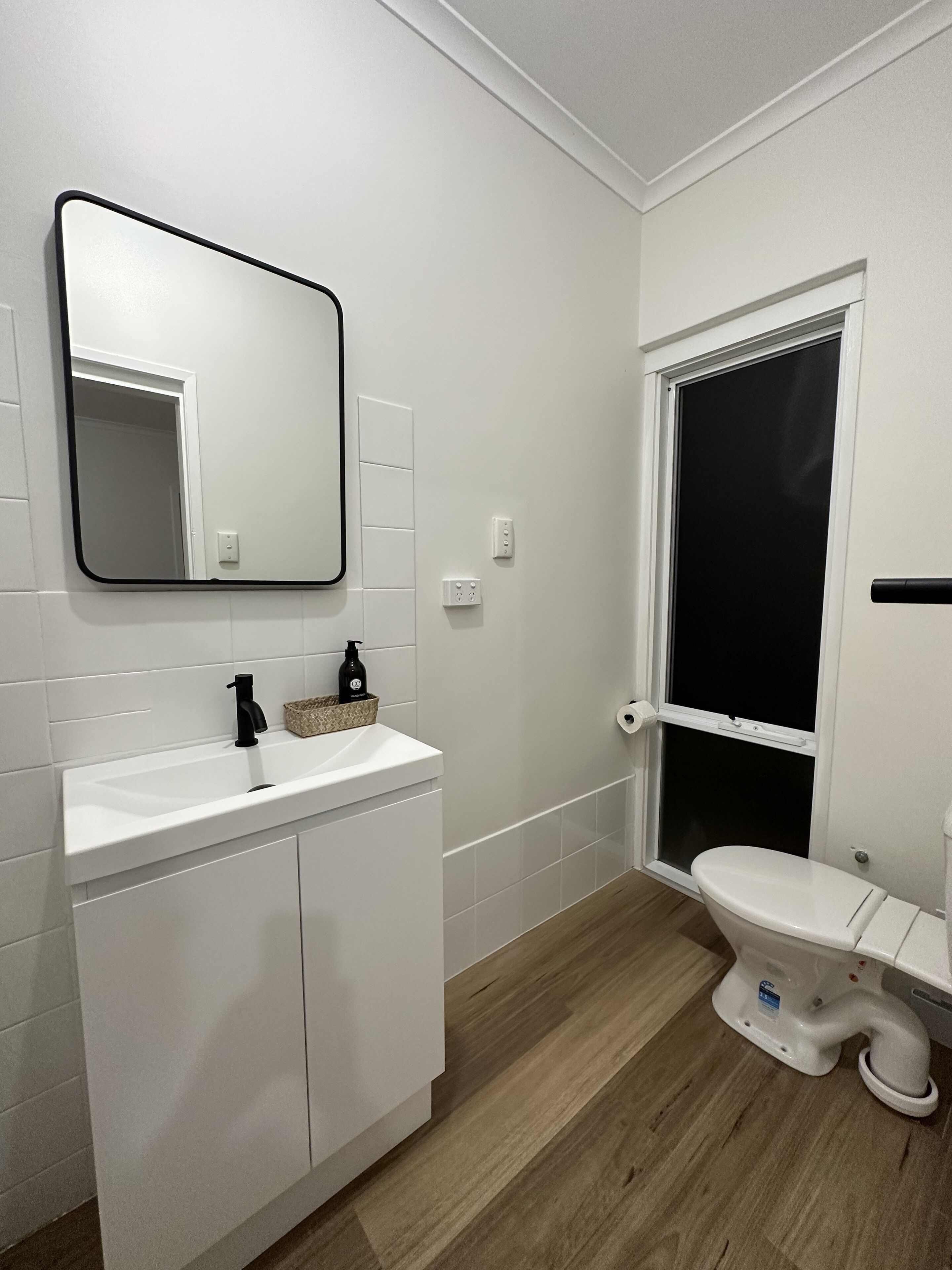 Ein modernes Badezimmer mit weißen Sanitärobjekten, Holzboden, Spiegel, Toilette und Fenster.