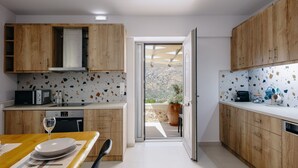 A modern kitchen with wooden cabinets, a dining table with a wine glass, and a view to an outdoor terrace. - Kedros Villa, A Luxury Touch (Rethymno)