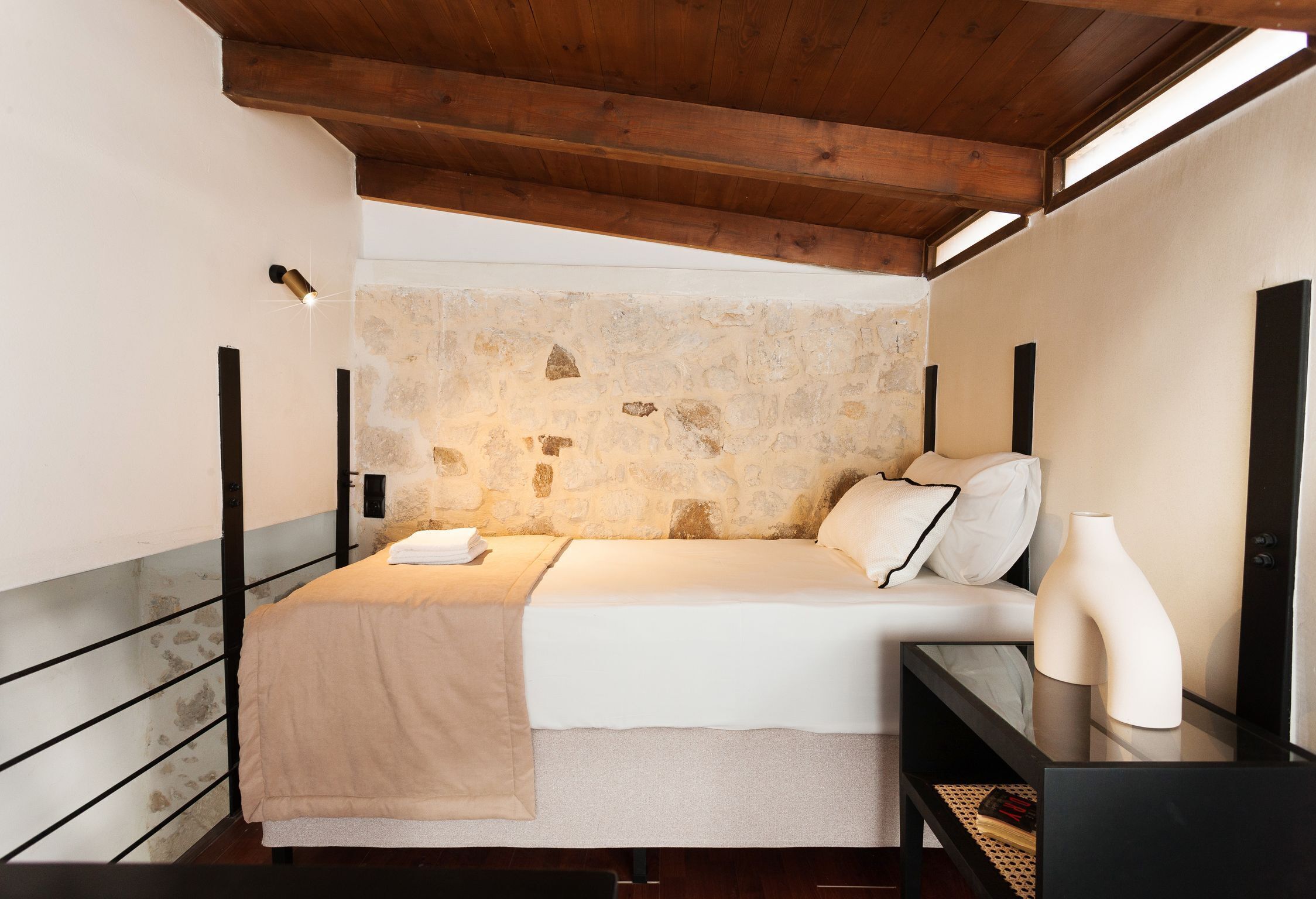 Ein Schlafzimmer mit Steinwand, Holzboden und einem Bett mit weißer Bettwäsche. Ein Nachttisch mit einer Vase und ein Beistelltisch mit einer Lampe.
