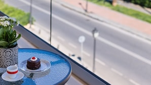 A balcony with a table, a potted plant, and a dessert. The view includes a marina with boats and a road with greenery and buildings in the background.