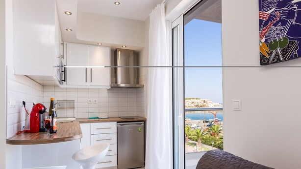 A modern kitchen with white cabinets, a stainless steel range hood, and a dishwasher. There is a dining area with a white table and two white chairs. A large window offers a view of a coastal area.