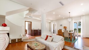 Uma sala de estar ampla com um sofá branco, uma mesa de centro e uma área de jantar com mesa e cadeiras de madeira.