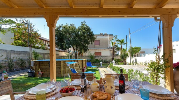 Een terras met een houten pergola, tafels gedekt met borden, glazen en een fles wijn.