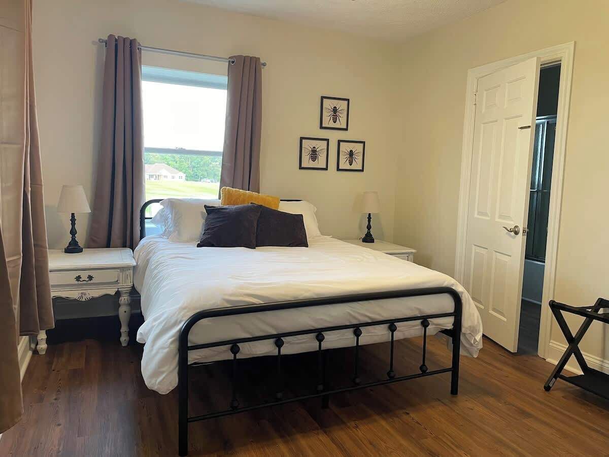 A bedroom with a bed, nightstands, a window with curtains, a door, and framed pictures on the wall.