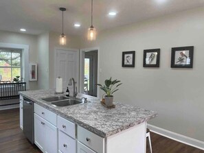 A kitchen with granite countertops, stainless steel appliances, pendant lighting, and framed artwork on the wall.
