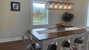 Uma sala de jantar com mesa de madeira, cadeiras brancas, luminária pendente e janela com vista.