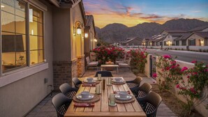 An outdoor dining area with a table set for four, surrounded by chairs, with a mountain backdrop and a sunset sky. - Mountain view townhouse w/ fire pit & pool (La Quinta)