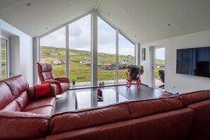 A spacious living room with a large window, a leather sofa, a red rocking chair, and a flat-screen TV mounted on the wall. - Luxurious Cabin | 3 Br | Scenic Village (Skalavik)