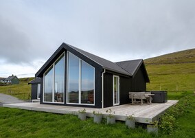 Ein modernes Haus mit großen Glasfenstern, einer Holzterrasse und einer grünen Landschaft.