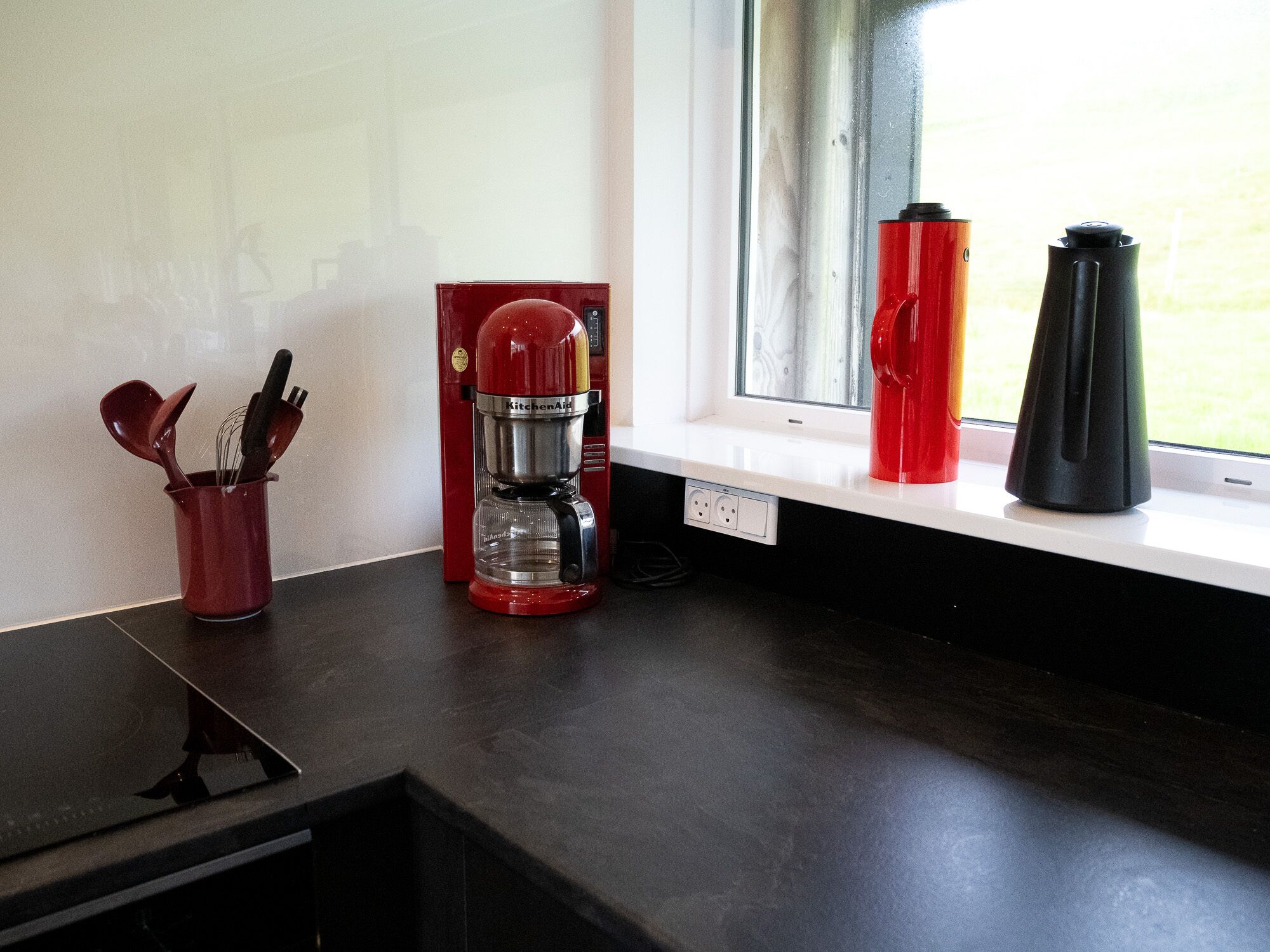 A kitchen counter with a red coffee maker, a red thermos, and a black coffee pot.