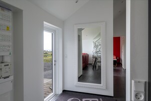 Ein Flur mit einem großen Spiegel, einer Tür zu einem Raum mit roter Wand und Blick auf eine ländliche Landschaft durch eine offene Tür.