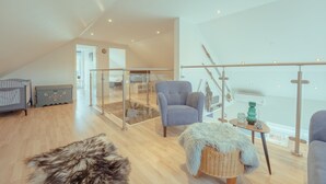 Een moderne woonkamer met een hoge plafond, houten vloer, een glazen scheidingswand, een grijze fauteuil, een rieten bijzettafeltje en een zacht tapijt.