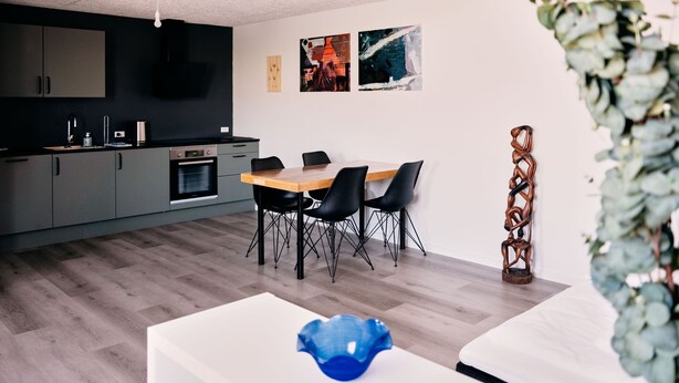 A modern kitchen with dark cabinets, a wooden dining table, and black chairs. There are two abstract paintings on the wall and a decorative plant on the right. - Studio Apartment | Unique Location | Stunning View (Hoyvík)