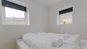 A bedroom with a large bed, two windows, and a view of a house and mountains. - Waterfront Retreat | 2 Br Boathouse (Oyrareingir)
