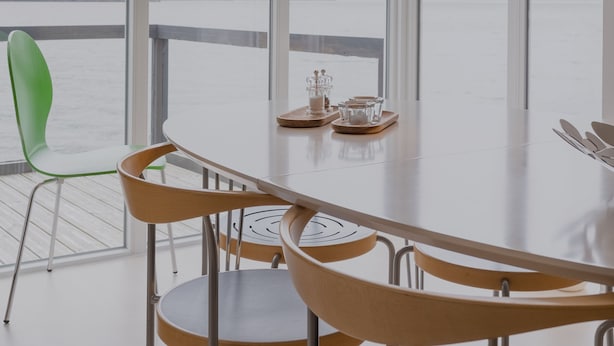 A modern dining area with a table and chairs, large windows with a view of a body of water and hills, and a unique textured pendant light. - Waterfront Retreat | 2 Br Boathouse (Oyrareingir)
