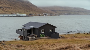 A modern house with a flat roof, a deck, and a view of a lake and mountains. - Waterfront Retreat | 2 Br Boathouse (Oyrareingir)
