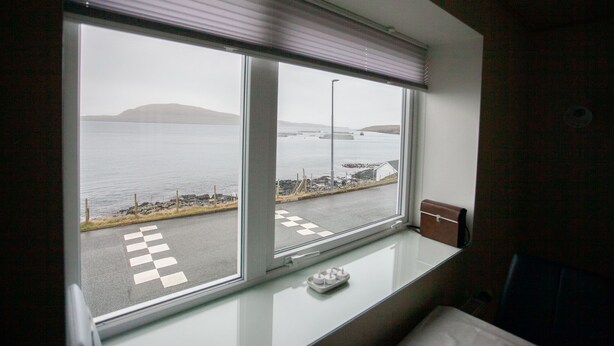 A room with a large window overlooking a coastal road with a checkered pattern, a small table, and a chair. - Charming 2Br Apartment, Sea View (Argir)