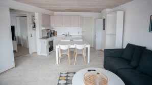 A modern kitchen with white cabinets, a dining table with chairs, and a dark-colored sofa in a living area. - Hyggeligt | Nordens Hus | Bager | Svartifossur (Tórshavn)