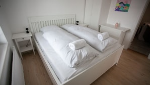 A white double bed with two pillows and a rolled towel, a bedside table with a lamp, and a small nightstand. - Hyggeligt | Nordens Hus | Bager | Svartifossur (Tórshavn)