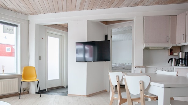 A modern living room with a wooden ceiling, a dining area with a white table and chairs, a kitchen with white cabinets and a black TV, and a yellow chair by a window. - Hyggeligt | Nordens Hus | Bager | Svartifossur (Tórshavn)
