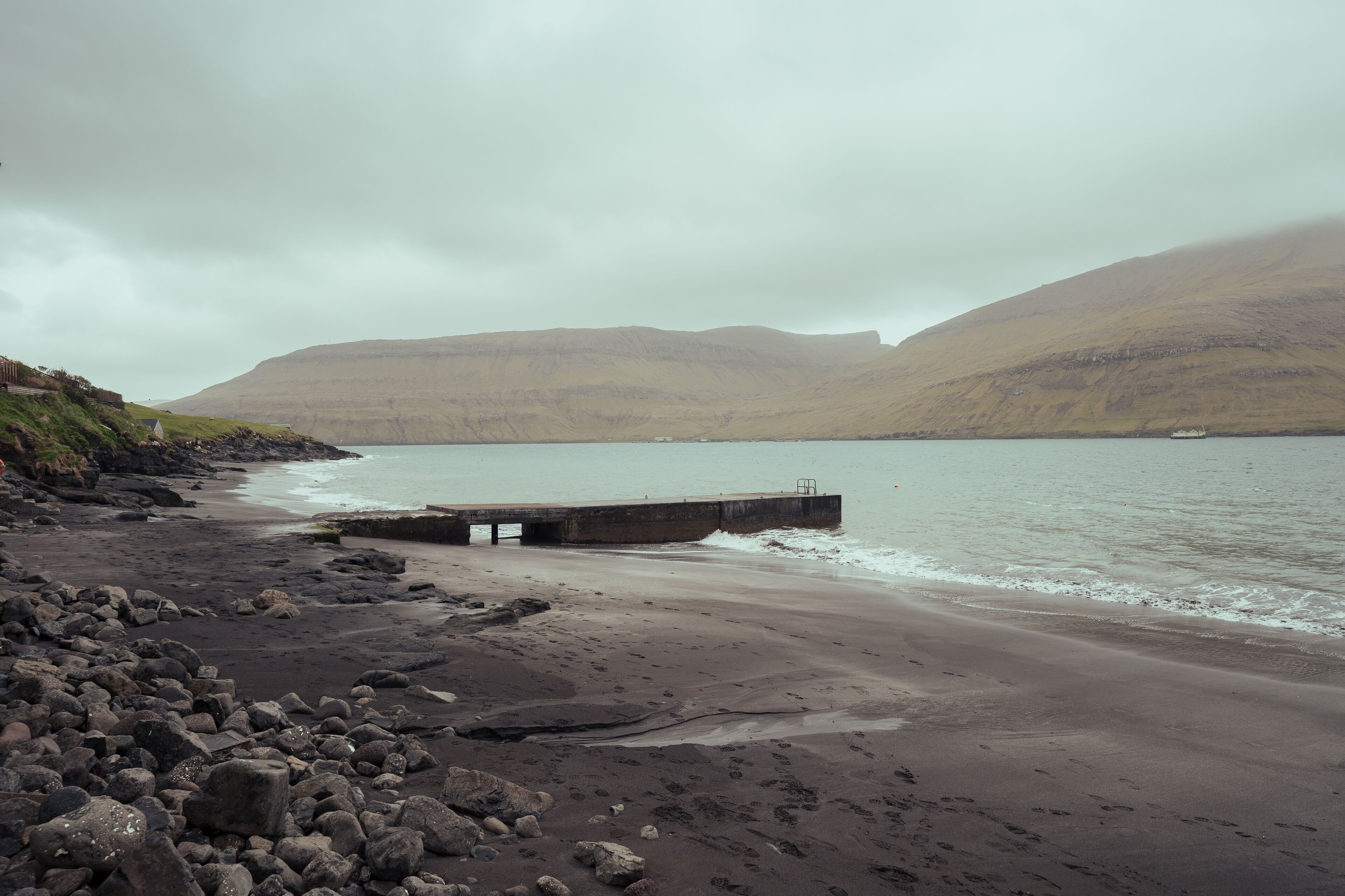 Une plage rocheuse avec une jetée qui s’avance dans des eaux calmes, le tout entouré de collines.