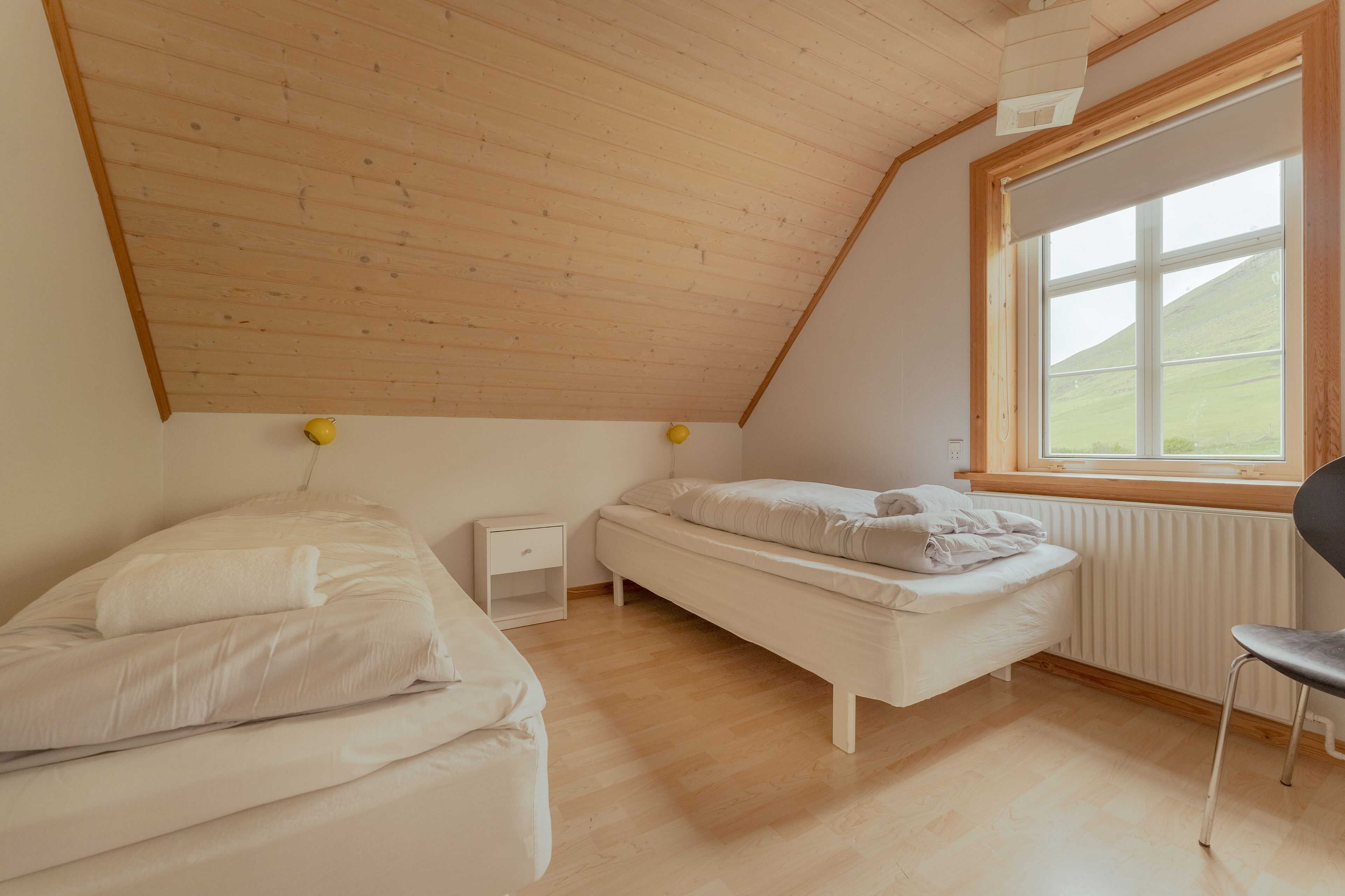 Un dormitorio con techo inclinado, ventana con vistas a un paisaje verde, cama, silla y mesita de noche.
