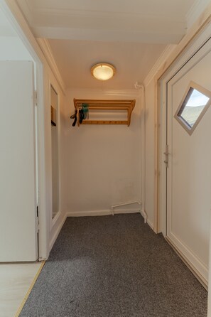 Un couloir avec un tapis gris, des murs blancs, une étagère en bois contenant des ustensiles de cuisine et un luminaire au plafond.
