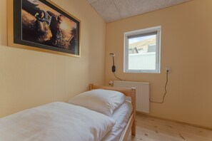 Un lit simple avec une literie blanche, dans une pièce aux murs de couleur beige, une fenêtre donnant sur des immeubles et un tableau accroché au mur.