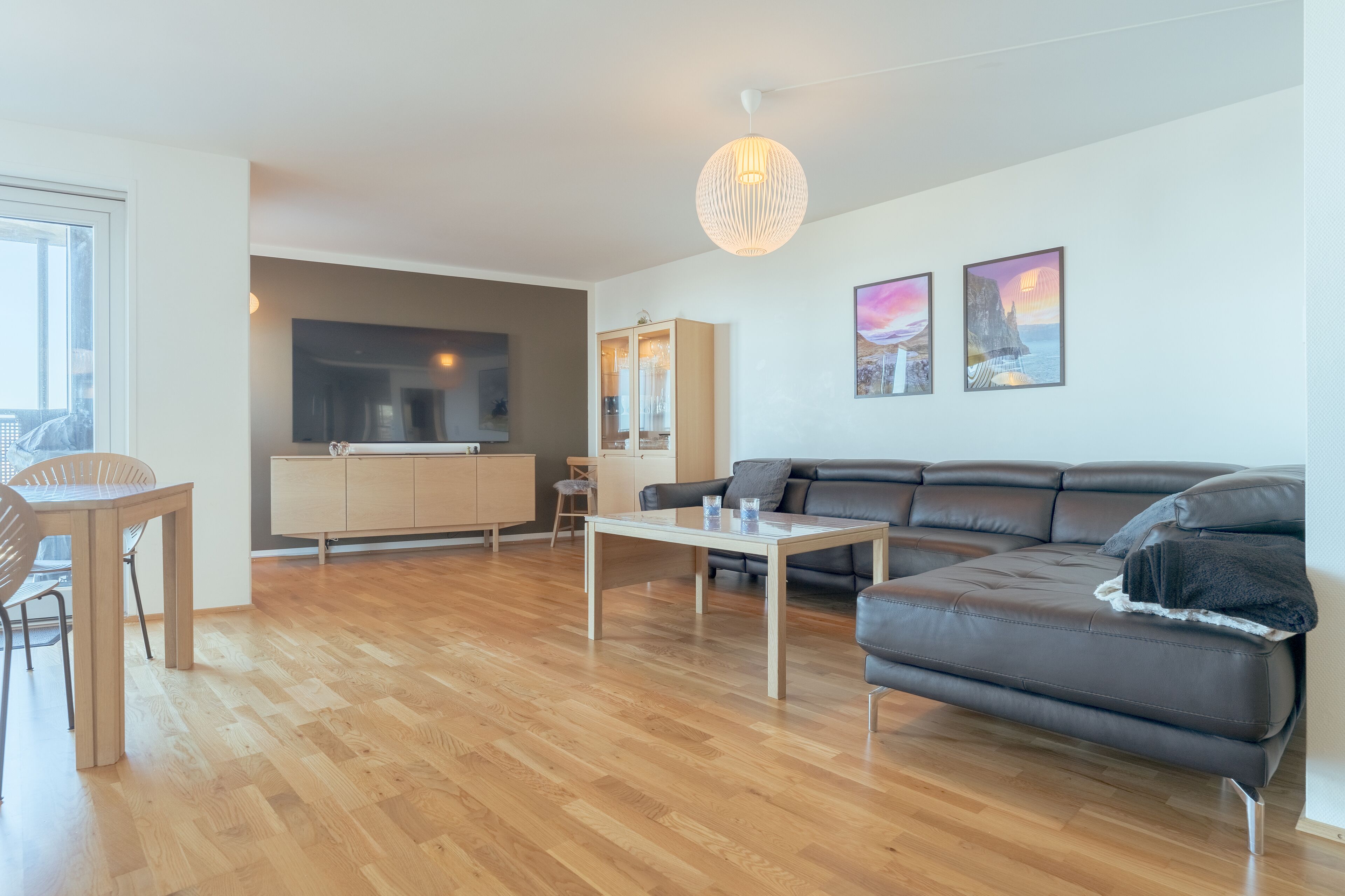A modern living room with a wooden floor, a black leather sofa, a white coffee table, and a dining area with a wooden table and chairs.