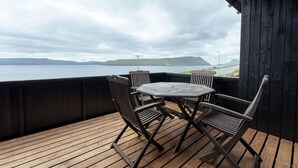 Une terrasse en bois avec une table et des chaises, d’où l’on peut admirer un plan d’eau et le paysage lointain.